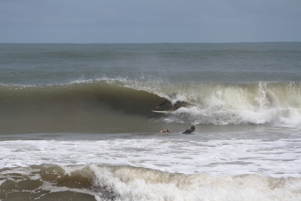 Surfista dentro do tubo em Regência num dia de mar cinza e revolto, enquanto outro rema para fora pela espuma